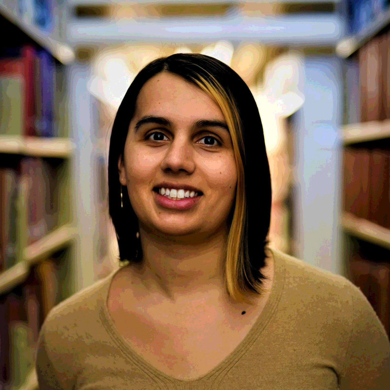 Image of Serena Lukas Bhandar, a smiling woman with brown hair and blonde highlights, wearing a tan dress, standing in a library.